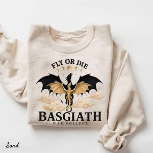 May include: Sand-colored sweatshirt featuring a dragon graphic with black and gold wings, clouds, and the text "FLY OR DIE" and "BASGIATH WAR COLLEGE". The design includes moons and stars, creating a fantasy theme.