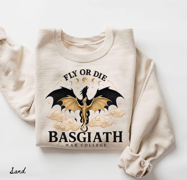May include: Sand-colored sweatshirt featuring a dragon graphic with black and gold wings, clouds, and the text "FLY OR DIE" and "BASGIATH WAR COLLEGE". The design includes moons and stars, creating a fantasy theme.