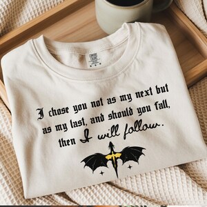May include: Cream-colored t-shirt with black gothic text: "I chose you not as my next but as my last, and should you fall, then I will follow." A black dragon graphic with yellow accents is below the text.
