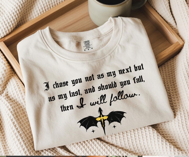 May include: Cream-colored t-shirt with black gothic text: "I chose you not as my next but as my last, and should you fall, then I will follow." A black dragon graphic with yellow accents is below the text.