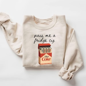 May include: Cream-colored sweatshirt with the phrase "pass me a fridge cig" in black script. A graphic of a Diet Coke cigarette box is below the text, with Diet Coke cans inside. The sweatshirt has rolled-up sleeves.