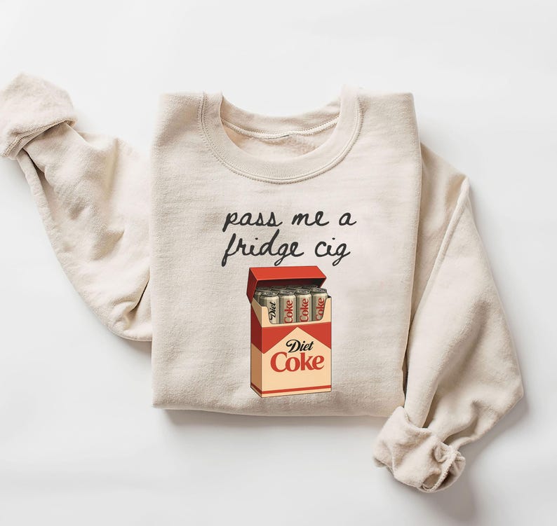 May include: Cream-colored sweatshirt with the phrase "pass me a fridge cig" in black script. A graphic of a Diet Coke cigarette box is below the text, with Diet Coke cans inside. The sweatshirt has rolled-up sleeves.