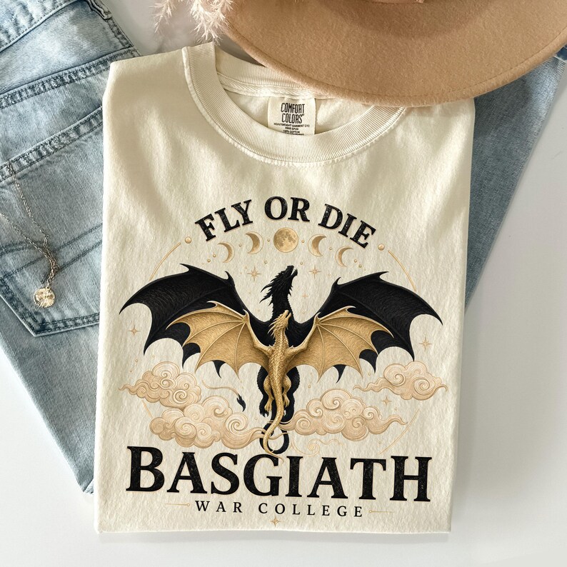 May include: Cream-coloured t-shirt featuring a dragon graphic and the text "FLY OR DIE" and "BASGIATH WAR COLLEGE". The dragon has black and gold wings. The shirt is made of a soft material.
