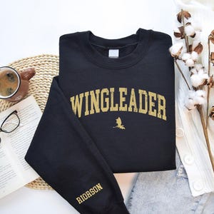 May include: Black sweatshirt with a gold graphic that says "WINGLEADER" with a small dragon silhouette underneath. The left sleeve has the word "RIORSON" in gold lettering.