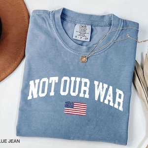 May include: Blue jean colored t-shirt with the words "NOT OUR WAR" in white block letters and a small American flag graphic. A gold necklace with a pendant rests on the shirt.