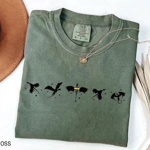 May include: A moss green t-shirt with a black graphic of five dragons flying in a row. The dragons are silhouetted against a white background with small white stars. The shirt has a tag that says "COMFORT COLORS" and "S - P - CH".