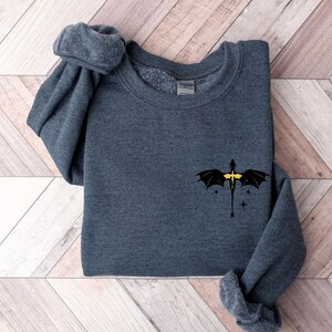 Puede incluir: Una sudadera gris oscuro con un dise&ntilde;o de drag&oacute;n negro en la parte delantera. El drag&oacute;n tiene detalles amarillos y est&aacute; volando con las alas extendidas. Hay peque&ntilde;as estrellas blancas alrededor del drag&oacute;n.