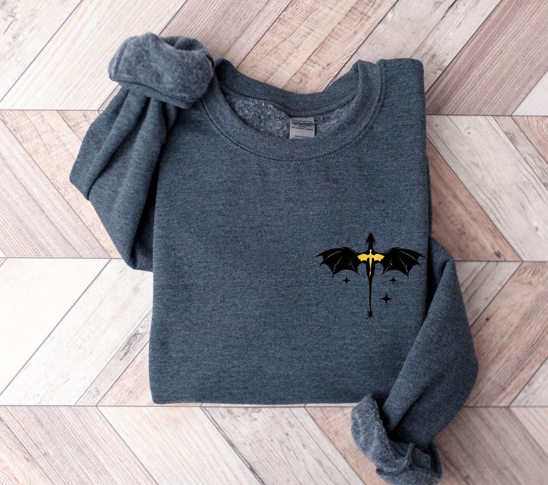 Puede incluir: Una sudadera gris oscuro con un dise&ntilde;o de drag&oacute;n negro en la parte delantera. El drag&oacute;n tiene detalles amarillos y est&aacute; volando con las alas extendidas. Hay peque&ntilde;as estrellas blancas alrededor del drag&oacute;n.