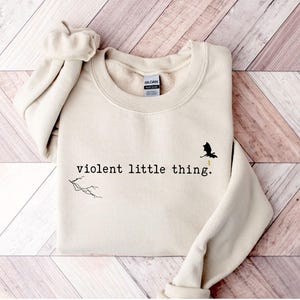 以下が含まれることがあります： クリーム色のスウェットシャツに、黒い文字で「violent little thing.」とプリントされています。テキストの上に、黄色い稲妻がついた小さな黒いドラゴンのシルエットがあります。