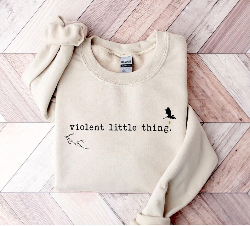 以下が含まれることがあります： クリーム色のスウェットシャツに、黒い文字で「violent little thing.」とプリントされています。テキストの上に、黄色い稲妻がついた小さな黒いドラゴンのシルエットがあります。