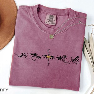 May include: Berry-colored t-shirt with a crew neck and a dragon graphic. The graphic features six stylized black dragons with a crescent moon and stars. A silver necklace with a pendant is draped across the shirt.