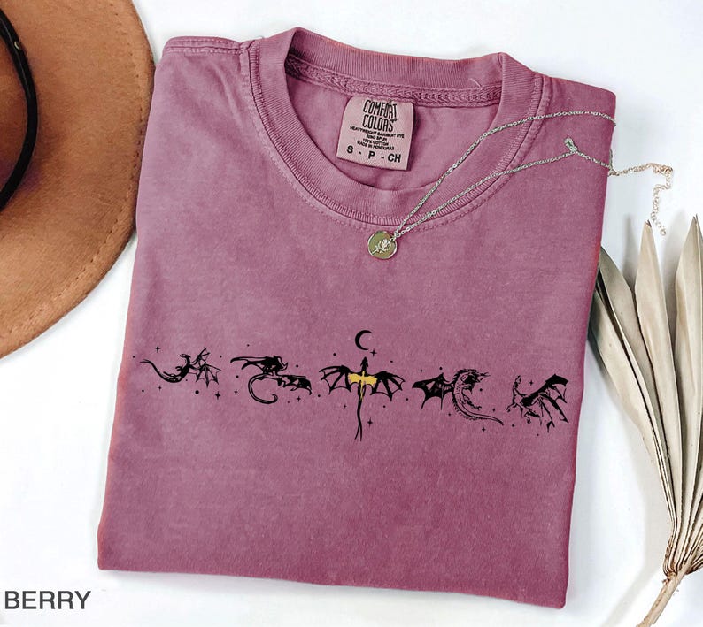 May include: Berry-colored t-shirt with a crew neck and a dragon graphic. The graphic features six stylized black dragons with a crescent moon and stars. A silver necklace with a pendant is draped across the shirt.
