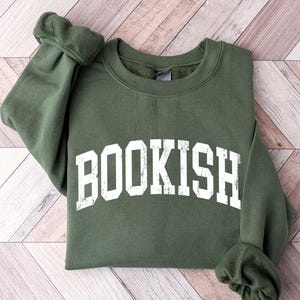 May include: An olive green sweatshirt with the word "BOOKISH" printed in large, distressed white letters across the front. The sweatshirt is folded, with the sleeves visible, and rests on a light-colored wooden surface.