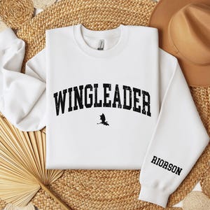 May include: White sweatshirt with black text that reads "WINGLEADER" in an arch with a small black silhouette of a bat underneath. The right sleeve has the word "RIORSON" printed in black.