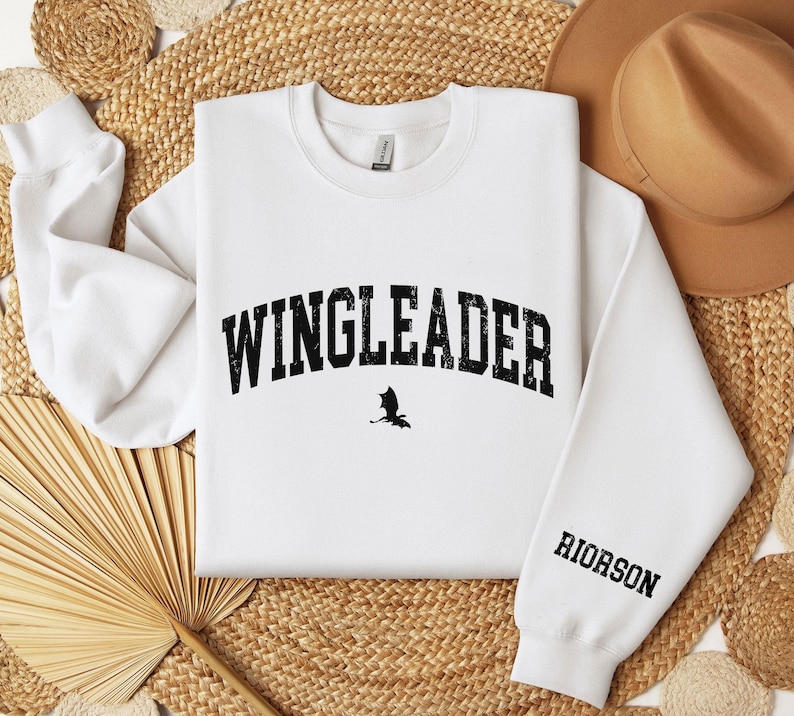 May include: White sweatshirt with black text that reads "WINGLEADER" in an arch with a small black silhouette of a bat underneath. The right sleeve has the word "RIORSON" printed in black.