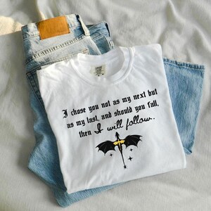 May include: White t-shirt with the quote "I chose you not as my next but as my last, and should you fall, then I will follow." and a dragon graphic. The shirt is laid on top of a pair of blue jeans.