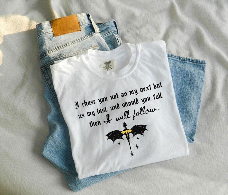 May include: White t-shirt with the quote "I chose you not as my next but as my last, and should you fall, then I will follow." and a dragon graphic. The shirt is laid on top of a pair of blue jeans.