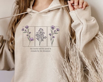 May include: A beige hoodie with a graphic of three floral sprigs in rectangular frames. The text "the world will be saved & remade by the dreamers" is printed below the graphic.