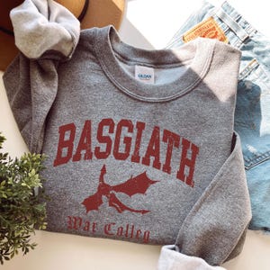 May include: A heather gray sweatshirt with the word "BASGIATH" in distressed red lettering, along with a red dragon graphic. The text "War Collen" is below the dragon. The sweatshirt is folded, with a gray scrunchie and denim jeans visible.