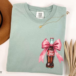 May include: A light green t-shirt with a pink bow and a Diet Coke bottle graphic. The bottle is red and white with the text "Diet Coke" printed on it.