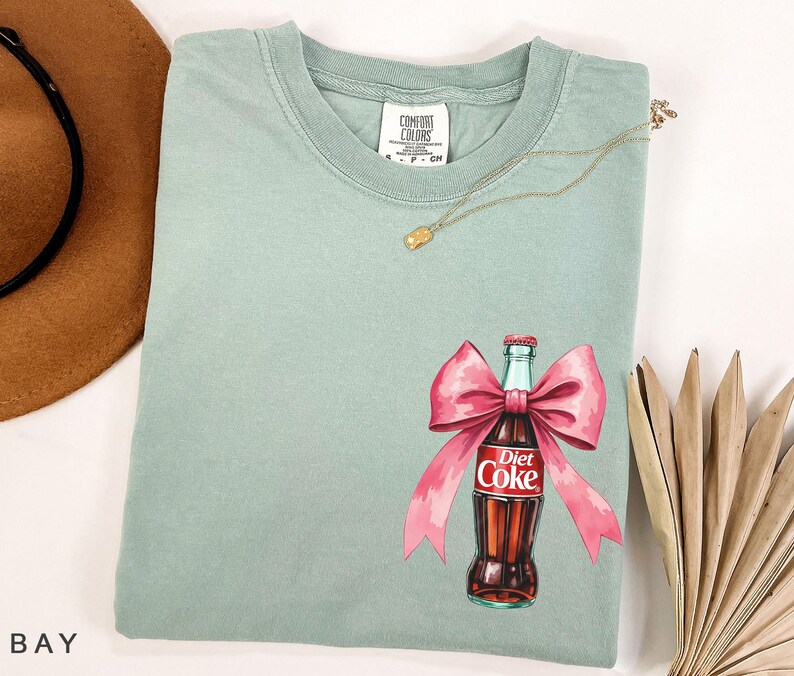 May include: A light green t-shirt with a pink bow and a Diet Coke bottle graphic. The bottle is red and white with the text "Diet Coke" printed on it.