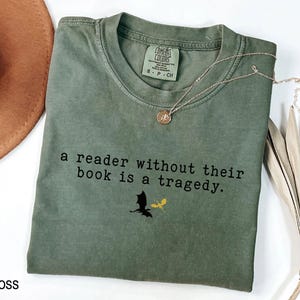 May include: Moss green t-shirt with the text "a reader without their book is a tragedy." A black dragon and a yellow dragon are printed below the text. A gold necklace with a pendant is on the shirt.