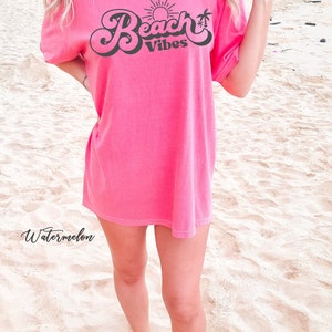 May include: A pink t-shirt with the text "Beach Vibes" in a retro font. The text is in black and features a sun and palm trees. The shirt is being worn by a person standing on a sandy beach.
