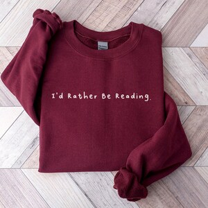K&ouml;nnte beinhalten: Ein weinroter Pullover mit dem Text "I'd Rather Be Reading." in Wei&szlig;.