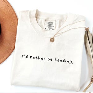 May include: A white t-shirt with the text "I'd Rather Be Reading." printed in black. The shirt is folded and laid flat on a white surface.