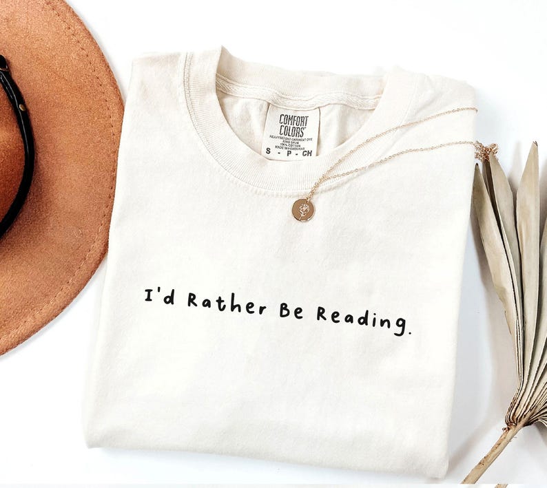 May include: A white t-shirt with the text "I'd Rather Be Reading." printed in black. The shirt is folded and laid flat on a white surface.