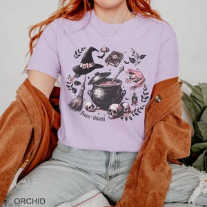 May include: A lavender t-shirt with a black and white illustration of a witch's cauldron, a broom, a frog, a skull, and a book. The text "Feelin' Witchy" is printed below the cauldron.