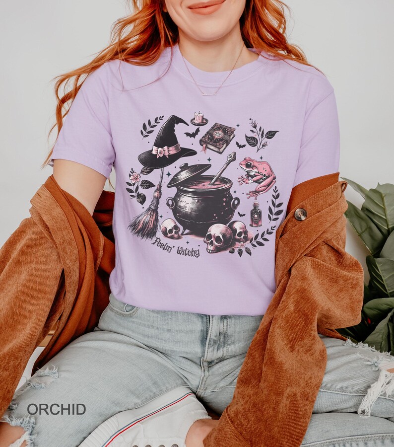 May include: A lavender t-shirt with a black and white illustration of a witch's cauldron, a broom, a frog, a skull, and a book. The text "Feelin' Witchy" is printed below the cauldron.