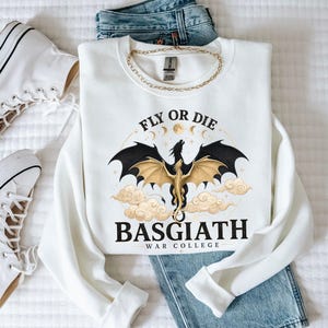 May include: White sweatshirt with a graphic of a dragon with black wings and gold accents, the text "FLY OR DIE" and "BASGIATH WAR COLLEGE". The sweatshirt is paired with blue jeans, a gold chain, and white sneakers.