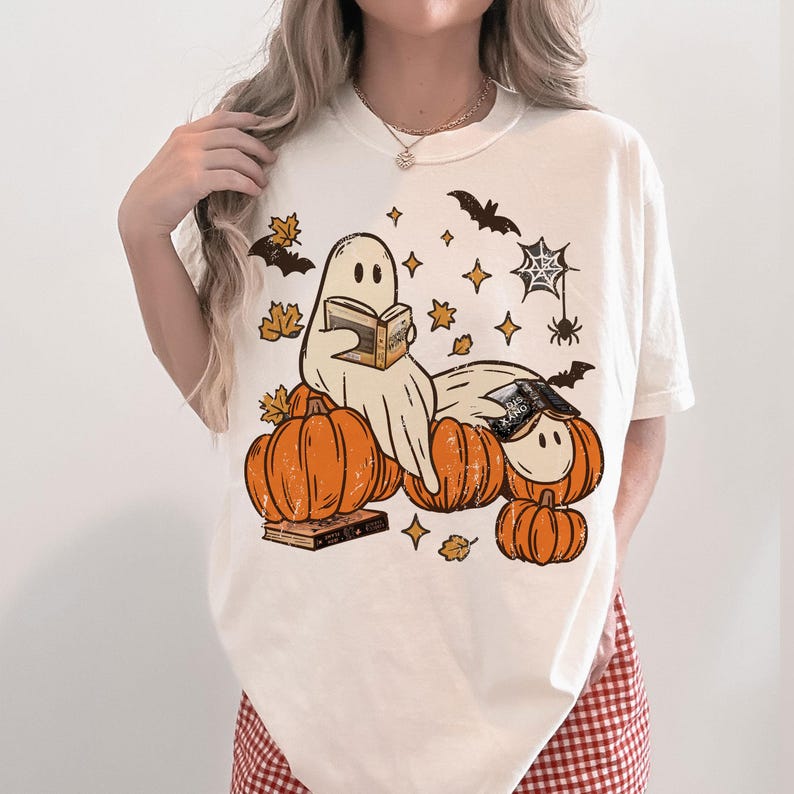 Może przedstawiać: Kremowy t-shirt z nadrukiem graficznym przedstawiającym dwa duszki czytające książki, siedzące wśr&oacute;d pomarańczowych dyń. Wz&oacute;r zawiera czarne nietoperze, pajęczynę z pająkiem oraz rozrzucone ż&oacute;łte jesienne liście i gwiazdki na przodzie.