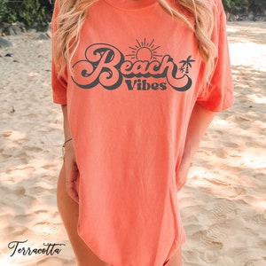 May include: A terracotta orange t-shirt with a retro-style graphic that says "Beach Vibes" in black. The graphic includes a sun and palm trees.