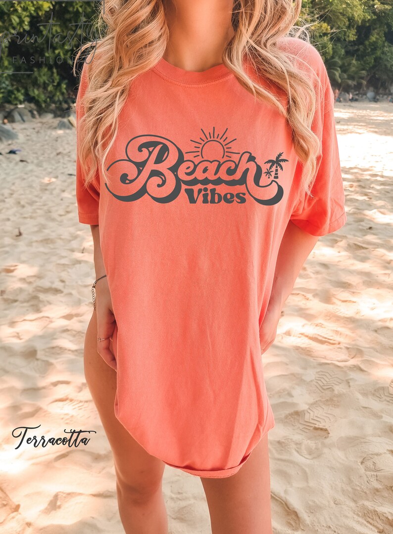 May include: A terracotta orange t-shirt with a retro-style graphic that says "Beach Vibes" in black. The graphic includes a sun and palm trees.