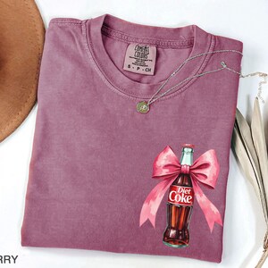 May include: A mauve colored t-shirt with a graphic of a Diet Coke bottle tied with a pink bow. The text "Diet Coke" is visible on the bottle.