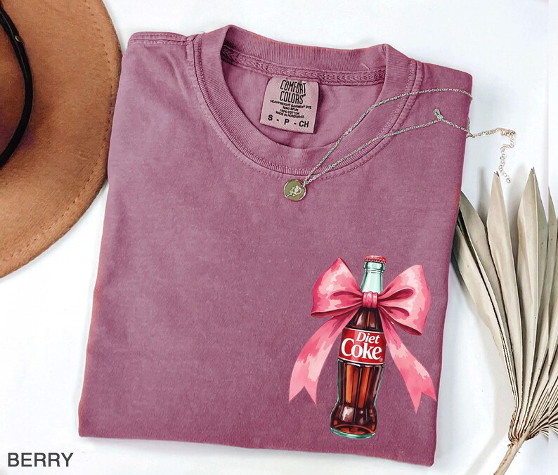 May include: A mauve colored t-shirt with a graphic of a Diet Coke bottle tied with a pink bow. The text "Diet Coke" is visible on the bottle.