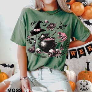 May include: Green t-shirt with a witchy graphic featuring a cauldron, a broom, a frog, a hat, and a book. The text on the shirt reads "Feelin' Witchy".