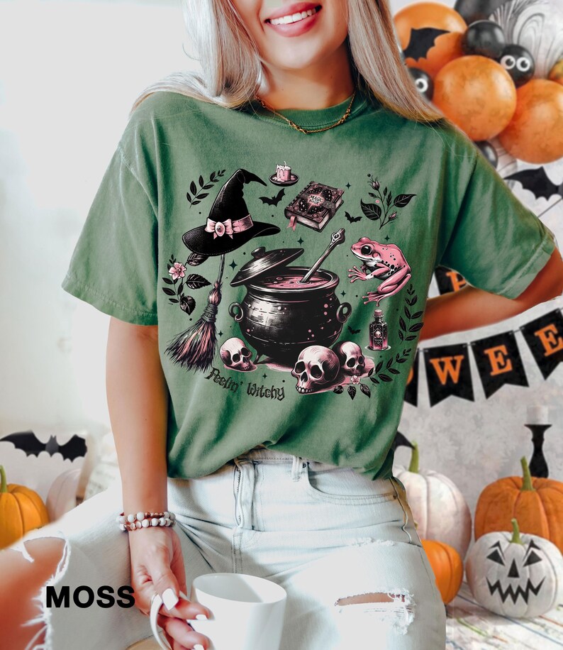 May include: Green t-shirt with a witchy graphic featuring a cauldron, a broom, a frog, a hat, and a book. The text on the shirt reads "Feelin' Witchy".