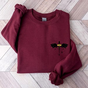 Puede incluir: Sudadera color burdeos con un gr&aacute;fico de drag&oacute;n negro y detalles amarillos. El drag&oacute;n est&aacute; volando con las alas extendidas y tiene una peque&ntilde;a estrella a cada lado.