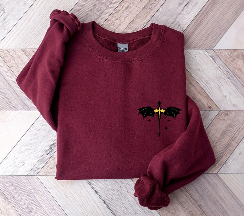 Puede incluir: Sudadera color burdeos con un gr&aacute;fico de drag&oacute;n negro y detalles amarillos. El drag&oacute;n est&aacute; volando con las alas extendidas y tiene una peque&ntilde;a estrella a cada lado.