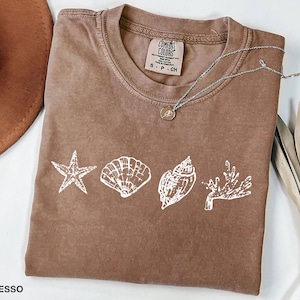 Op de afbeelding: Een bruin T-shirt met een witte afbeelding van schelpen en een zeester. Het shirt heeft de tekst "COMFORT COLORS" en "S - P - CH" gedrukt op het label. Het shirt ligt plat op een wit oppervlak.