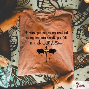 May include: A peach-colored t-shirt with the quote "I chose you not as my next but as my last, and should you fall, then I will follow." A black dragon graphic is centered below the text.