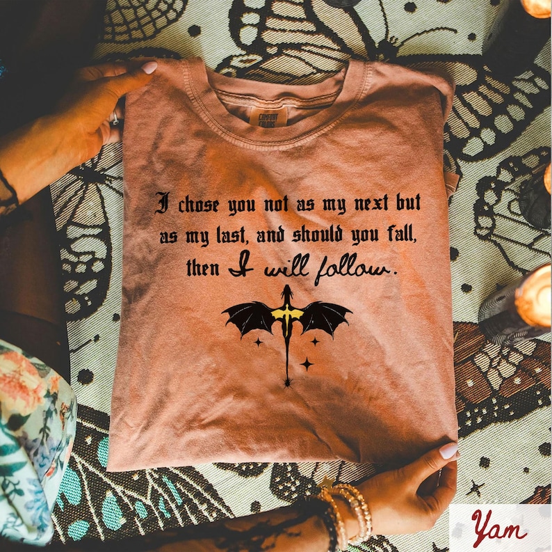 May include: A peach-colored t-shirt with the quote "I chose you not as my next but as my last, and should you fall, then I will follow." A black dragon graphic is centered below the text.