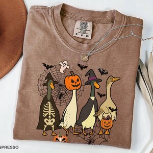 Puede incluir: Camiseta Comfort Colors color espresso con un gr&aacute;fico tem&aacute;tico de Halloween. El dise&ntilde;o incluye cuatro patos de dibujos animados disfrazados para Halloween, incluyendo un esqueleto, una bruja y una calabaza. Un collar plateado est&aacute; sobre la camiseta.