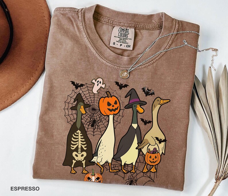 Puede incluir: Camiseta Comfort Colors color espresso con un gr&aacute;fico tem&aacute;tico de Halloween. El dise&ntilde;o incluye cuatro patos de dibujos animados disfrazados para Halloween, incluyendo un esqueleto, una bruja y una calabaza. Un collar plateado est&aacute; sobre la camiseta.