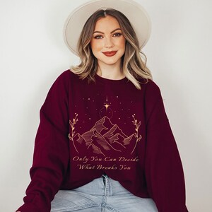 May include: Burgundy sweatshirt with a gold outline of mountains, stars, and a quote that reads "Only You Can Decide What Breaks You."