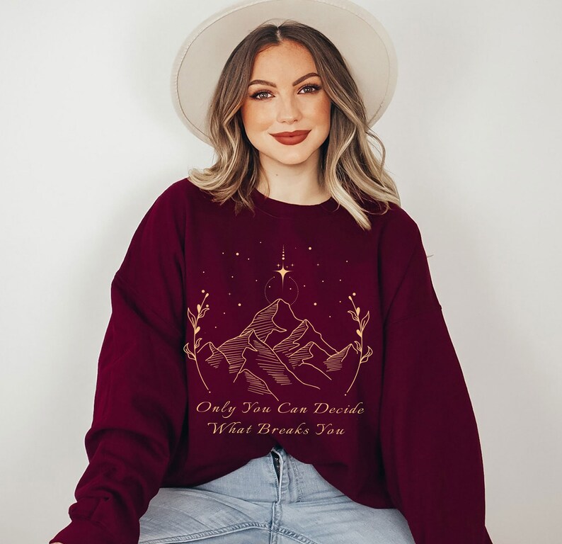 May include: Burgundy sweatshirt with a gold outline of mountains, stars, and a quote that reads "Only You Can Decide What Breaks You."