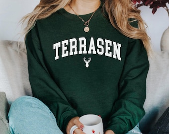 May include: A dark green sweatshirt with the word "TERRASEN" in white letters and a white deer silhouette. The sweatshirt is worn by a person sitting on a couch.